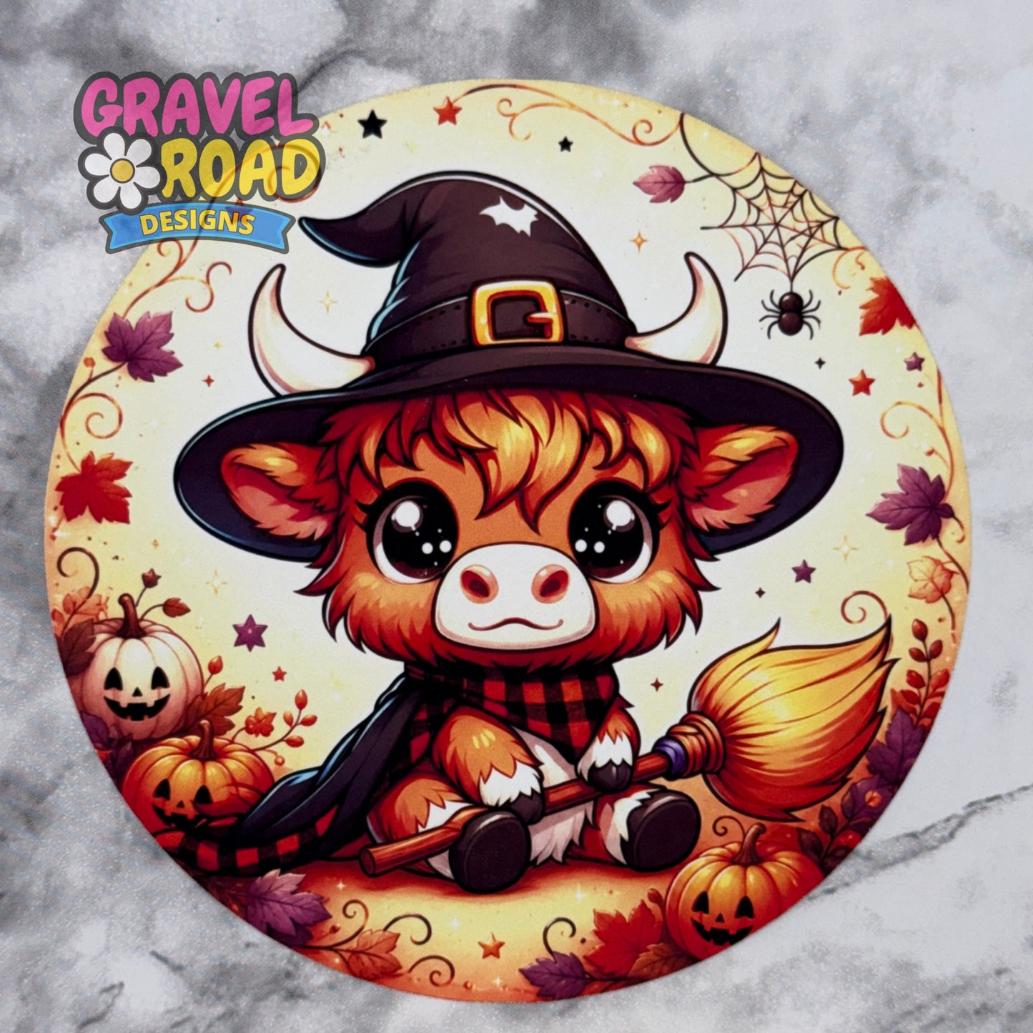 A circular cardstock featuring a cute cartoon Highland cow dressed as a witch, holding a broomstick and surrounded by pumpkins, autumn leaves, and spiderwebs. The brand logo 'Gravel Road Designs' is visible in the top left corner.