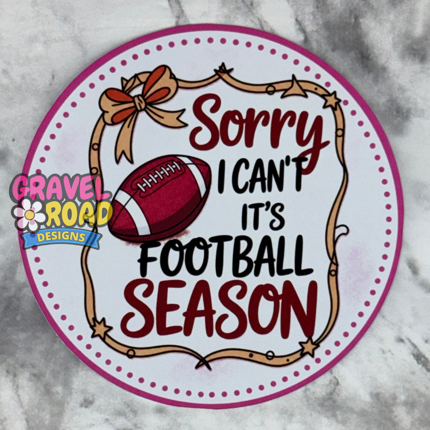 A round cardstock featuring a football with the words "Sorry I Can't It's Football Season". With a football and bow. On a marble-patterned surface, with a 'Gravel Road Designs' logo at the bottom.