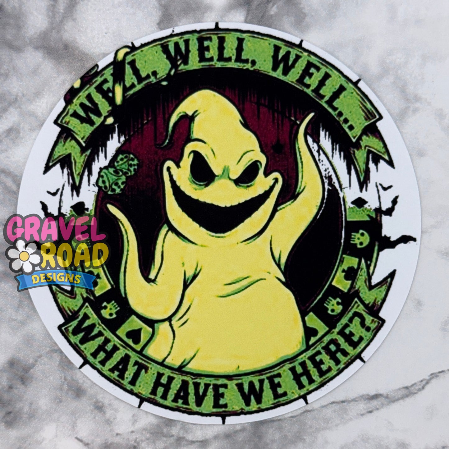 A circular cardstock featuring a yellow ghost with the text "WELL WELL WELL WHAT HAVE WE HERE?" The logo Gravel Road Designs on the left-hand side. On a grey marble backdrop.
