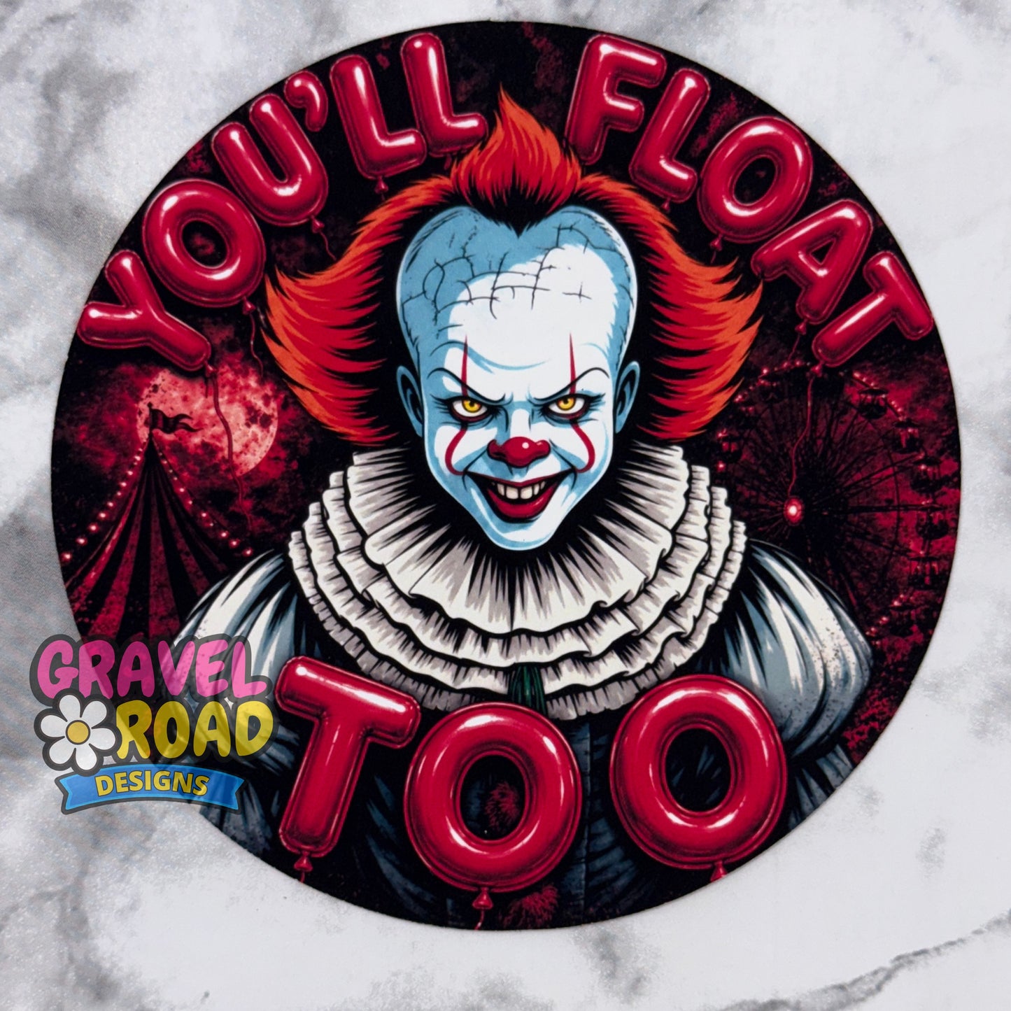 A circular cardstock design featuring a creepy clown illustration with red hair, blue face paint, and a white ruffled collar, surrounded by the text 'YOU'LL FLOAT TOO' in large red letters. A 'Gravel Road Designs' logo is visible in the lower-left corner. On a grey marble background.