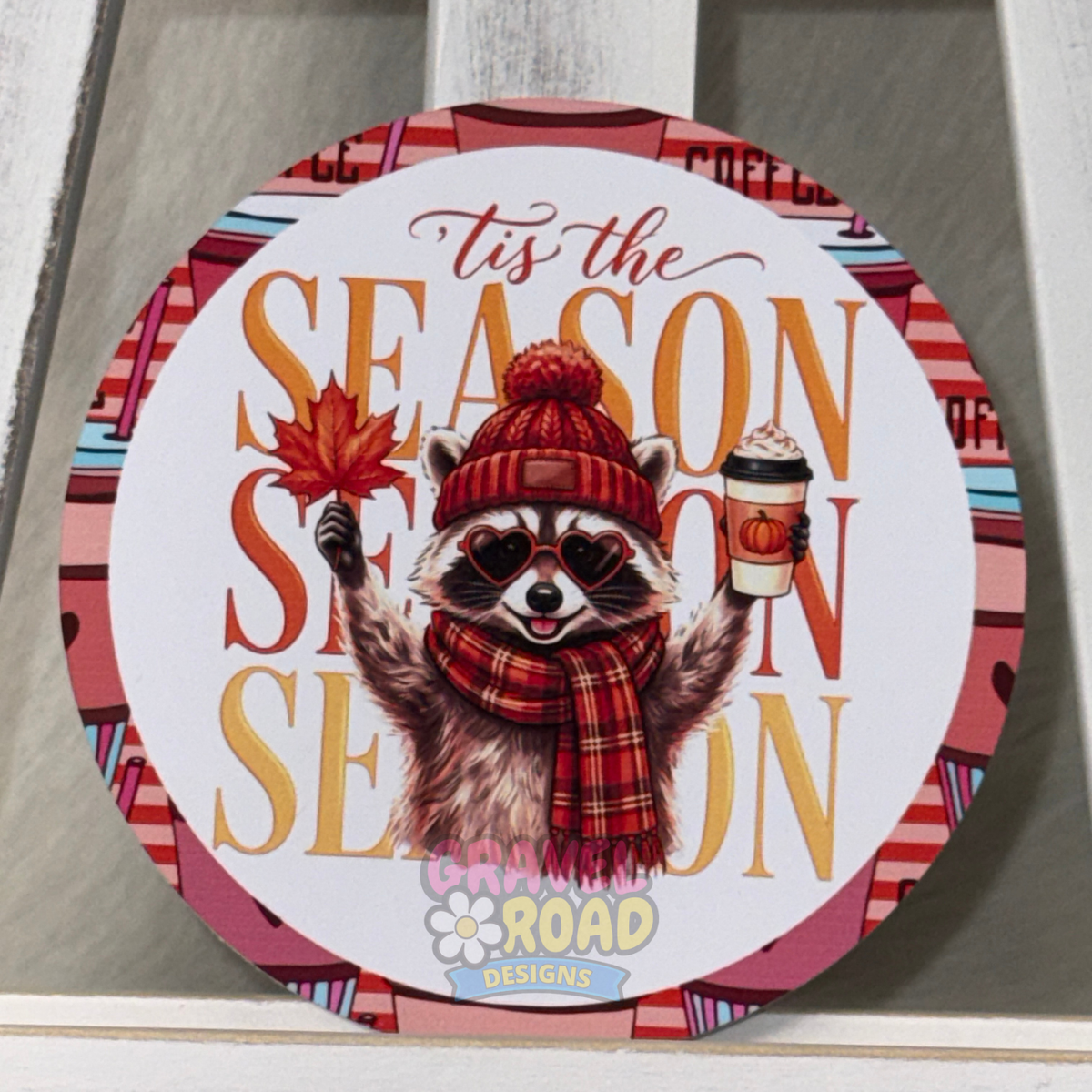 Round fall cardstock featuring a raccoon in heart-shaped sunglasses, a red beanie and plaid scarf, holding a leaf and pumpkin coffee. Text says "Tis The Season"