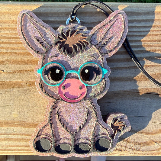 A glittery cartoon donkey-shaped car freshie with turquoise glasses and a pink snout, shown on a wooden surface with a black cord attached.