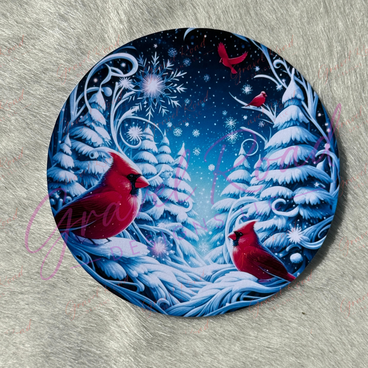 A round cardstock featuring a winter forest scene with two red cardinals, snow-covered pine trees, and a dark blue night sky, displayed on a gray textured surface.