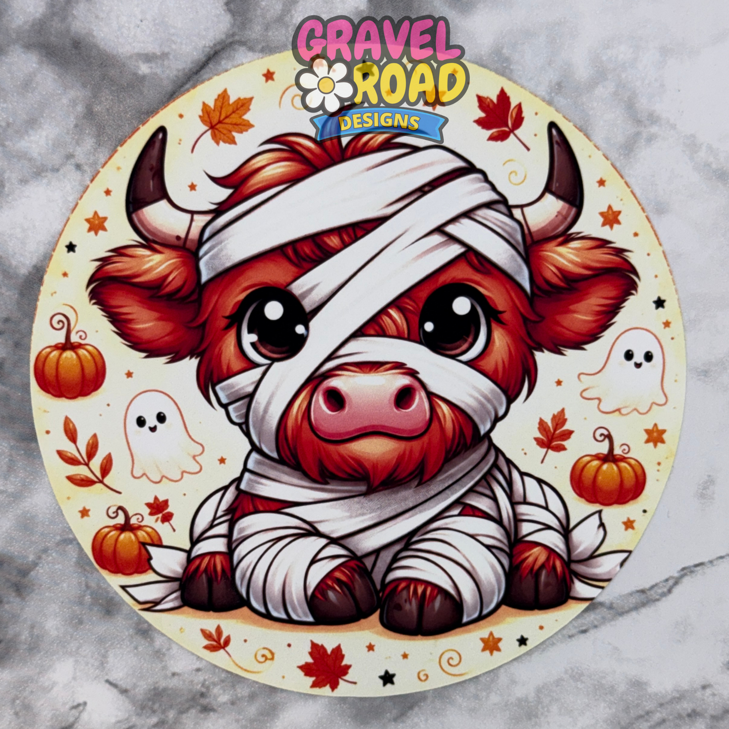A circular cardstock design featuring a cute cartoon mummy highland cow surrounded by Halloween elements like pumpkins, ghosts, and autumn leaves, with the brand logo 'Gravel Road Designs' at the top, displayed on a gray marble background.