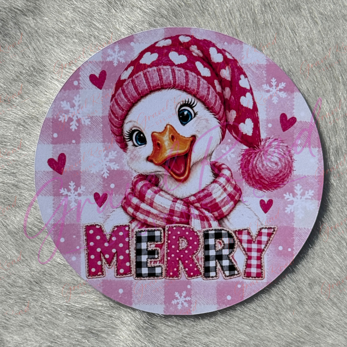 A round cardstock featuring a cute cartoon duck wearing a pink and white heart-patterned knit hat and matching scarf, with the word “MERRY” across the bottom on a pink gingham background with snowflakes and hearts. Displayed on a faux fur background.