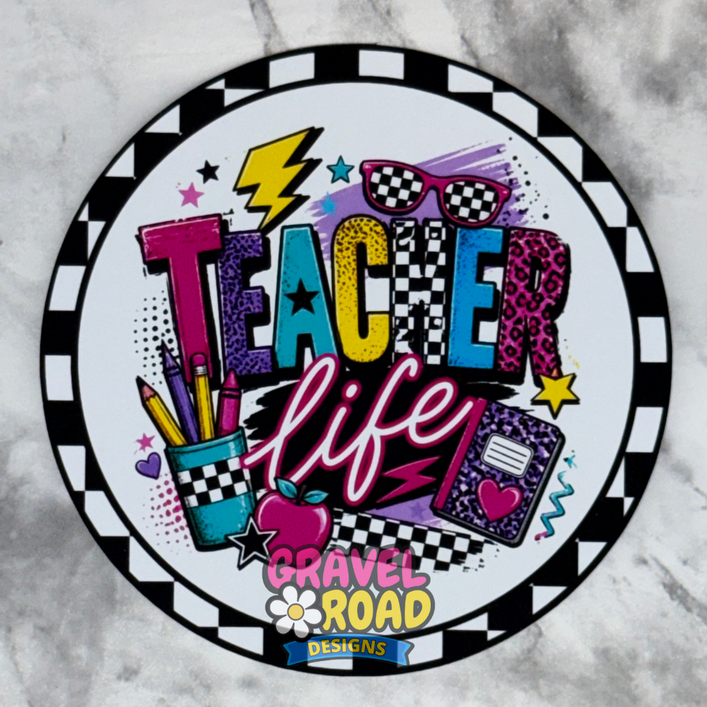 A circular cardstock design featuring the words 'Teacher Life' in colorful retro-style lettering, surrounded by school-themed doodles like pencils, an apple, sunglasses, and a notebook. The design includes a black-and-white checkered border and a 'Gravel Road Designs' logo at the bottom, shown on a light gray marble surface.
