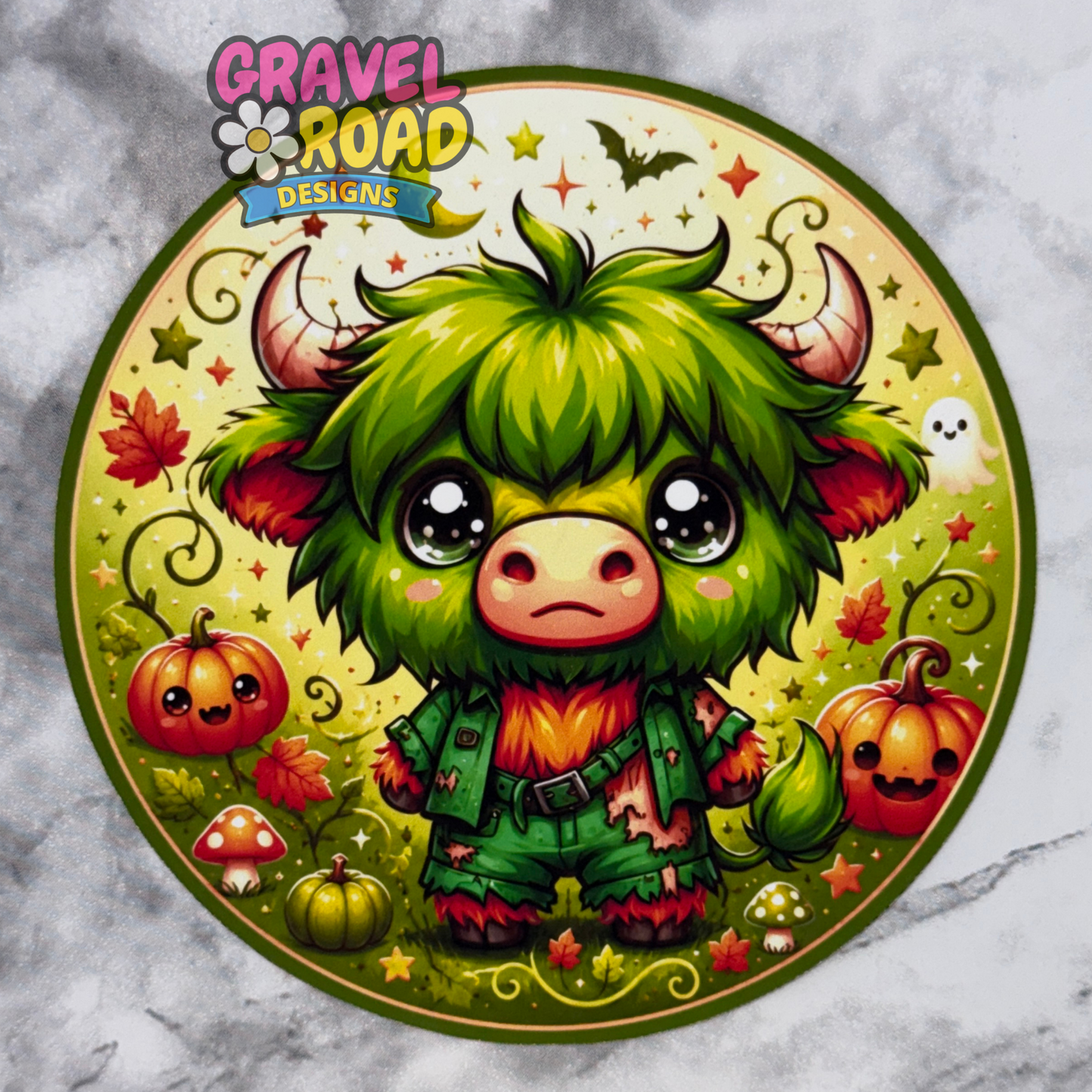 Round Halloween fall cardstock design featuring a cute green furry monster cow with big eyes, horns, and a patched green outfit. The background is yellow-green with pumpkins, mushrooms, fall leaves, stars, swirls, a small ghost, and a bat. A Gravel Road Designs watermark appears at the top left.