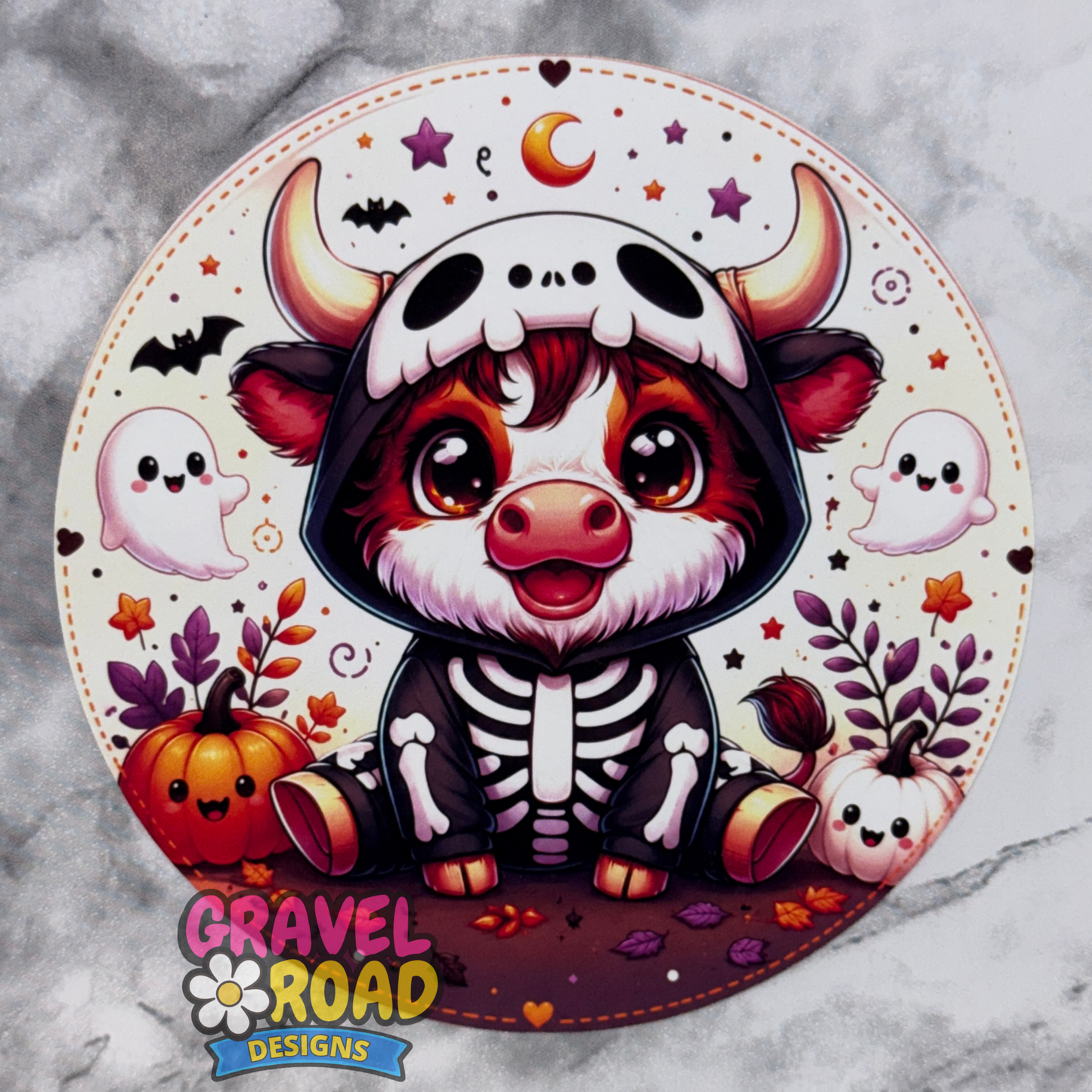 Round Halloween cow freshie cardstock design featuring a cute baby cow dressed in a black skeleton costume with a skull hood. The background has a cream and purple Halloween theme with smiling ghosts, bats, stars, pumpkins, fall leaves, and a crescent moon. A Gravel Road Designs watermark appears near the bottom left.