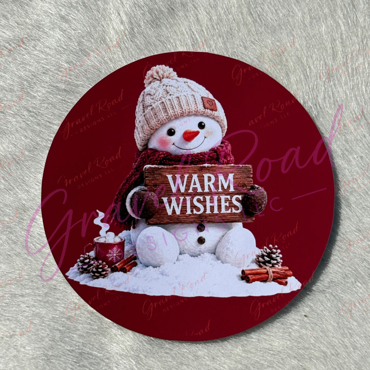A round cardstock featuring a festive snowman illustration. The snowman wears a cream knit hat and a red scarf, holding a wooden sign that reads 'WARM WISHES' in white letters.