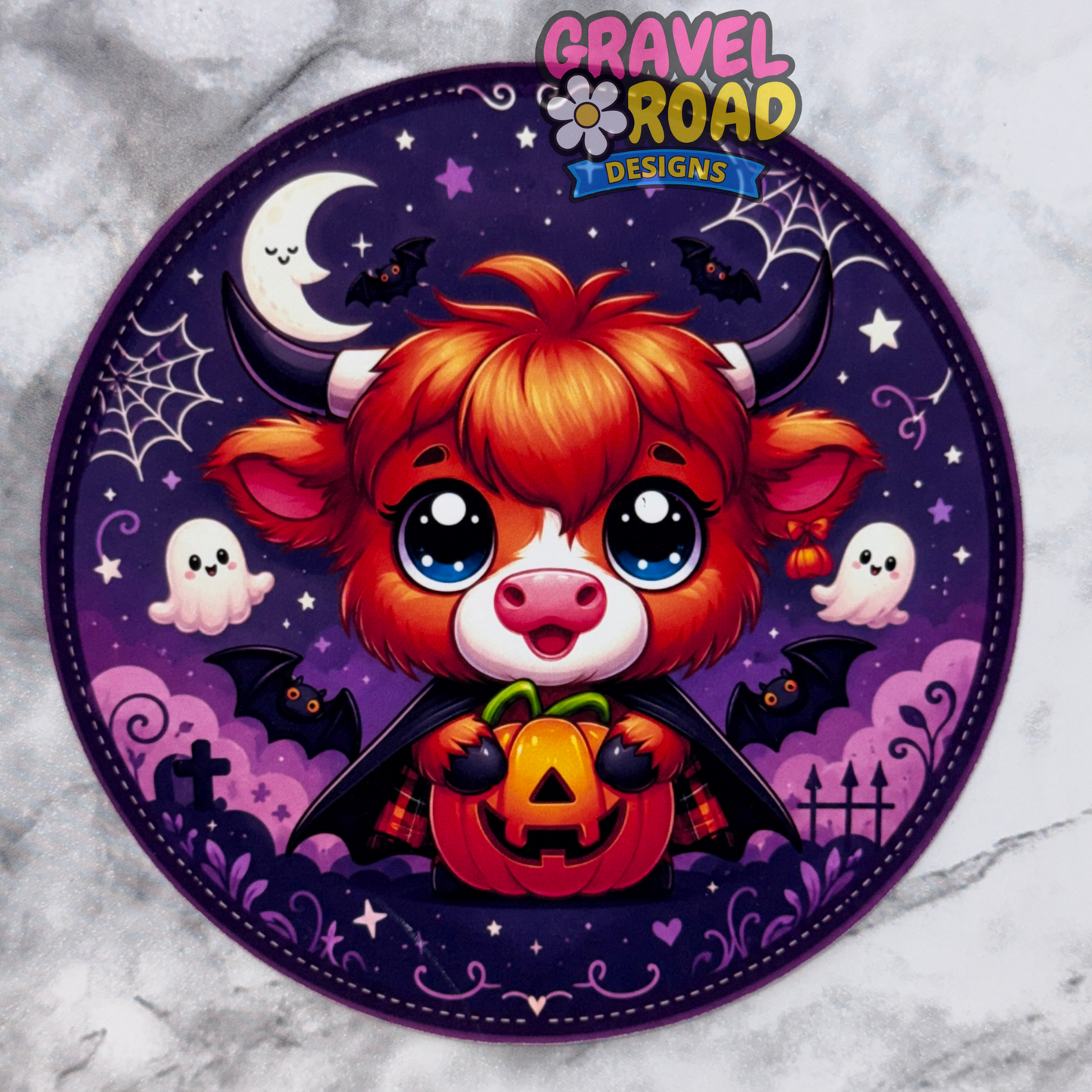 A circular Halloween-themed design on cardstock featuring a cute orange and white cartoon cow with large blue eyes, holding a jack-o'-lantern, set against a purple night sky with bats, ghosts, stars, and a crescent moon. The design includes the 'Gravel Road Designs' logo at the top and is displayed on a light gray marble background.
