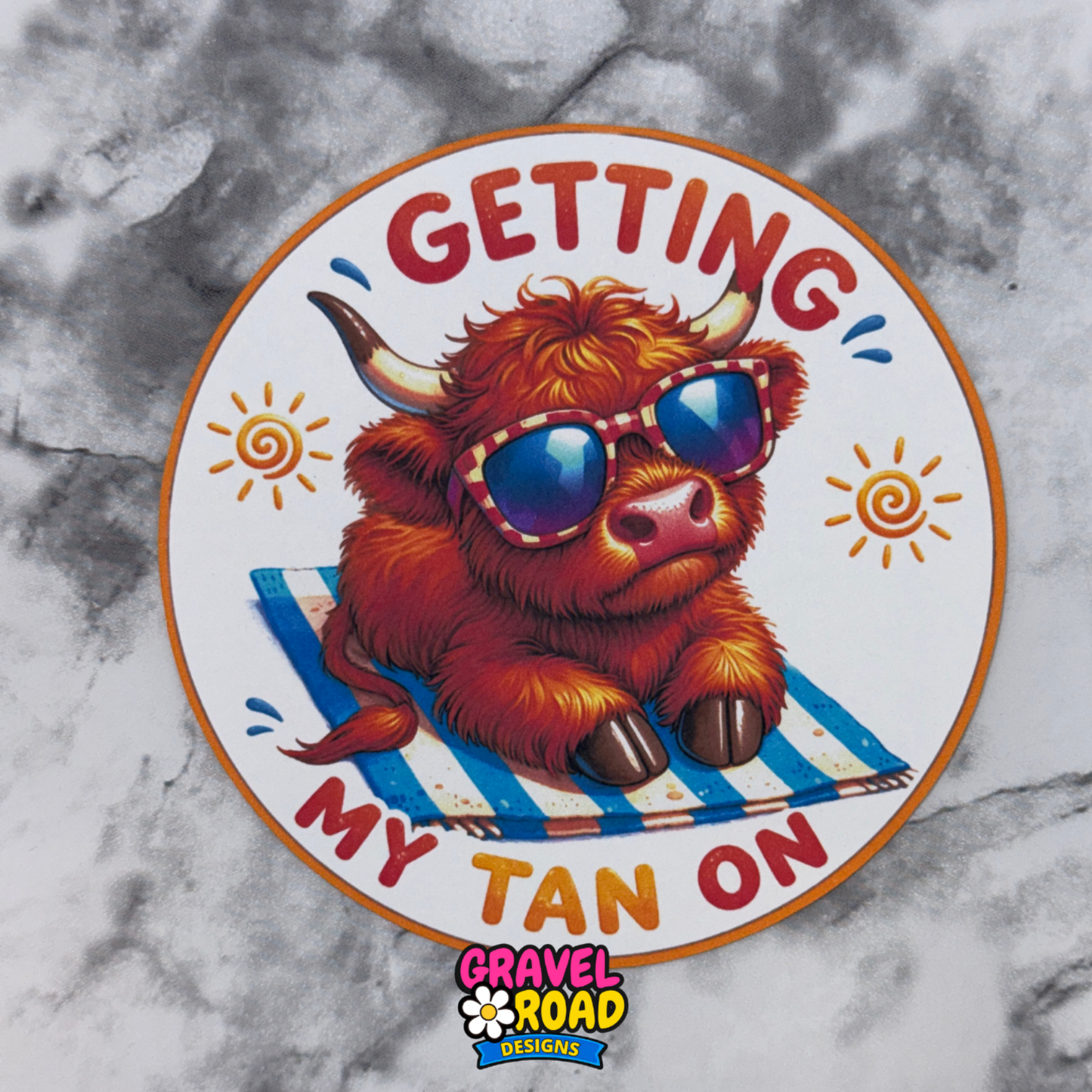Cardstock with a cartoon highland cow wearing sunglasses on a towel, text 'Getting My Tan On', and Gravel Road Designs logo on a marble background.