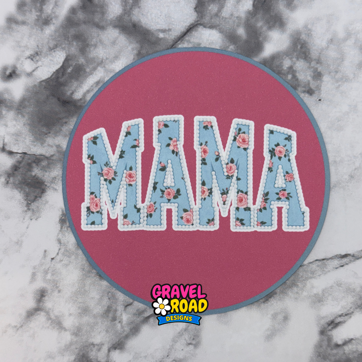 Circular cardstock printed in pink with the word "Mama" in blue with flowers. Gravel Road Designs logo printed on the bottom.