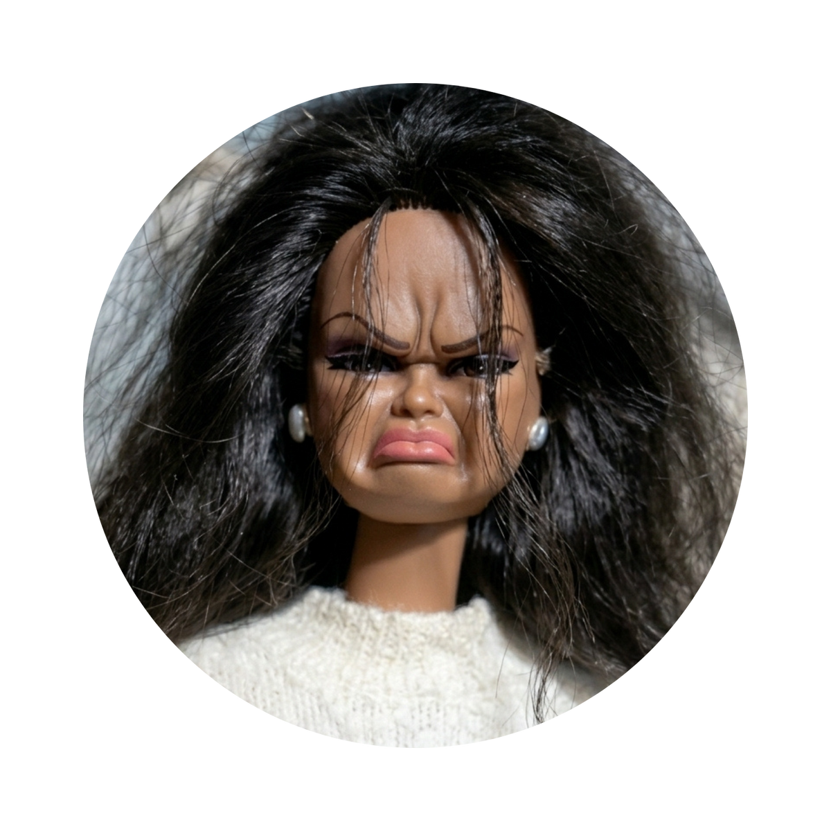 A close-up circular photo of a doll with a distressed, angry expression, wearing a cream knit top and small pearl-like earrings, isolated on a black background.