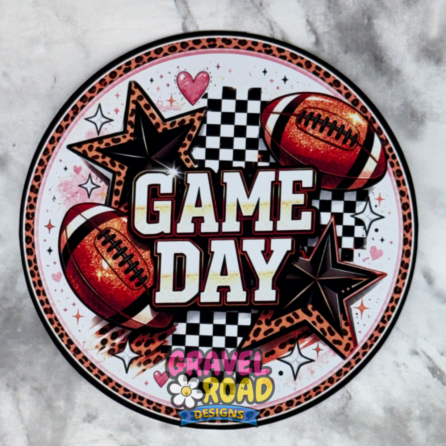 A circular cardstock featuring the words 'GAME DAY' in bold white letters with black outlines, surrounded by two footballs, black stars, checkered flags, and leopard-print accents. The design includes a 'Gravel Road Designs' logo at the bottom and is displayed on a gray marble background.