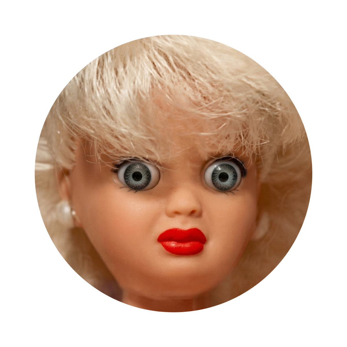 A close-up circular photo of a vintage blonde doll or collectible figure with large gray-blue eyes, red lips, and light pink skin, isolated on a black background.