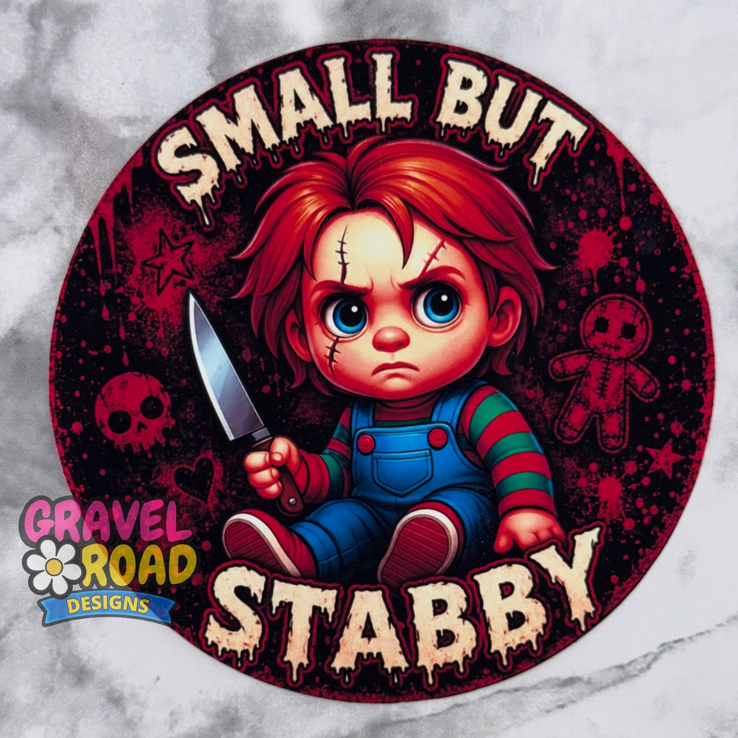 A circular cardstock design featuring a cartoon horror character with red hair, blue overalls, and a knife, surrounded by the text 'SMALL BUT STABBY' in dripping cream lettering. A 'Gravel Road Designs' logo appears in the lower-left corner. On a grey marble backdrop.