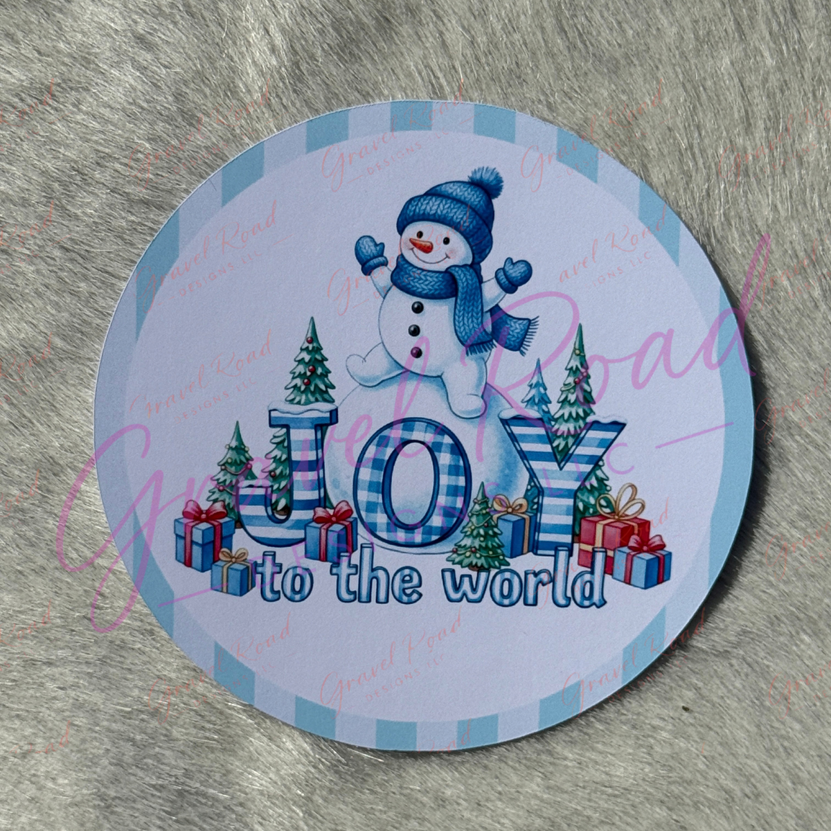 A round cardstock featuring a winter holiday design with a snowman, the text 'JOY to the world,' Christmas trees, and wrapped gifts on a light blue and white plaid border, placed on a gray faux fur surface.