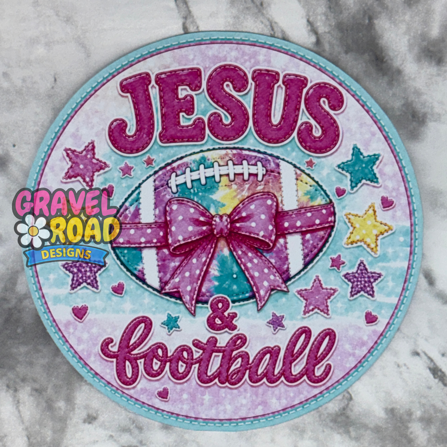 A round cardstock featuring the words 'JESUS & football' in glittery pink lettering, surrounding a football with a pink polka-dot bow. The patch is decorated with pink and blue stars and hearts on a light blue and pink background, with a 'Gravel Road Designs' logo on the left side.
