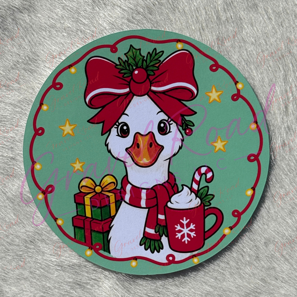 A round Christmas-themed cardstock featuring a cartoon white goose wearing a large red bow and striped scarf, holding a mug with whipped cream and a candy cane, next to wrapped gifts, on a mint green background with a red decorative border. Displayed on a faux fur background.