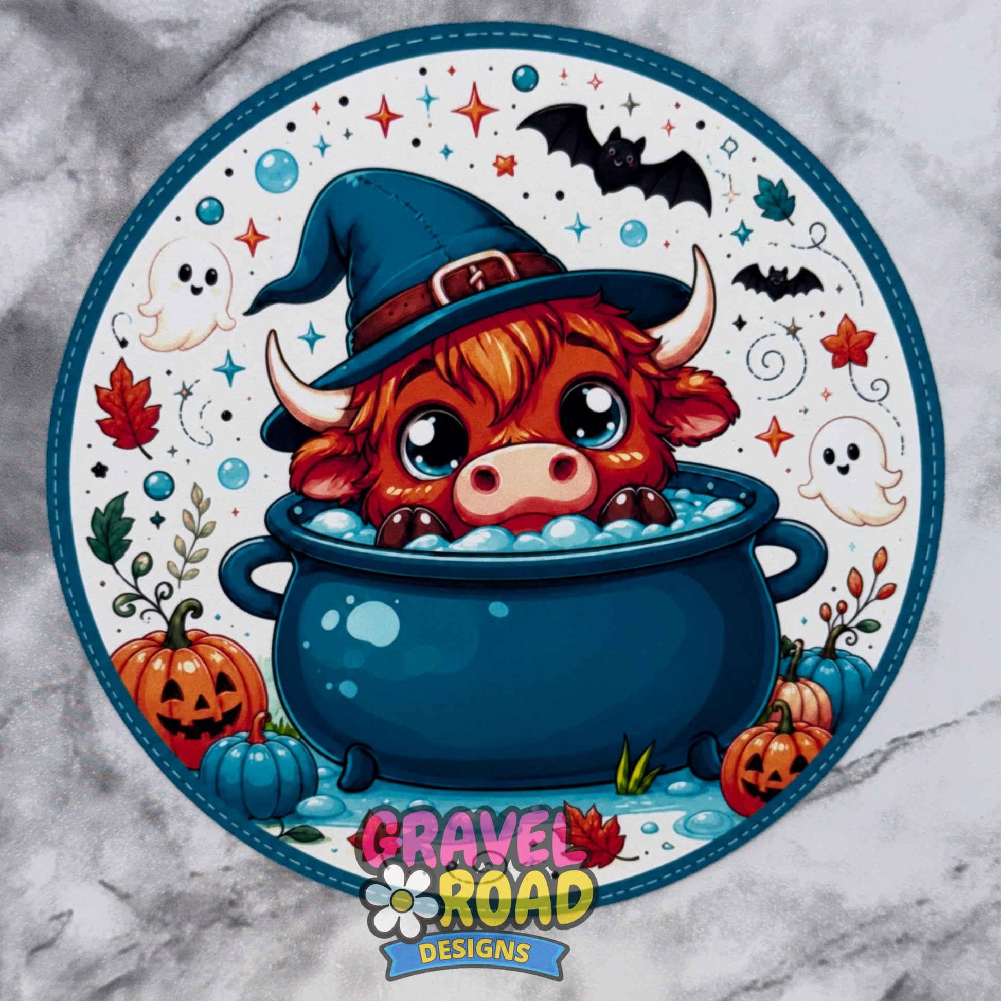 A circular Halloween-themed graphic cardstock featuring a cute red highland cow in a blue witch hat sitting in a cauldron with bubbles, surrounded by bats, ghosts, pumpkins, stars, and the logo 'GRAVEL ROAD DESIGNS' at the bottom, shown on a gray marble-like background.