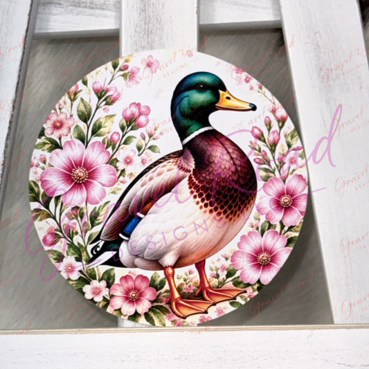 A round cardstock featuring a detailed illustration of a mallard duck surrounded by pink flowers, displayed on a white wooden surface.
