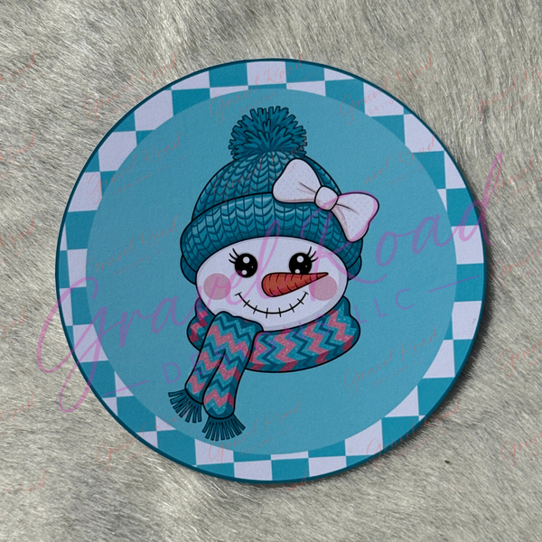A round printed cardstock featuring a cartoon snowman wearing a blue knit hat with a white bow and a blue-and-pink zigzag scarf, set against a teal and white checkered border. The item is photographed on a gray faux-fur surface.