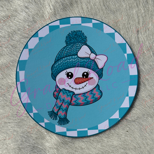 A round printed cardstock featuring a cartoon snowman wearing a blue knit hat with a white bow and a blue-and-pink zigzag scarf, set against a teal and white checkered border. The item is photographed on a gray faux-fur surface.