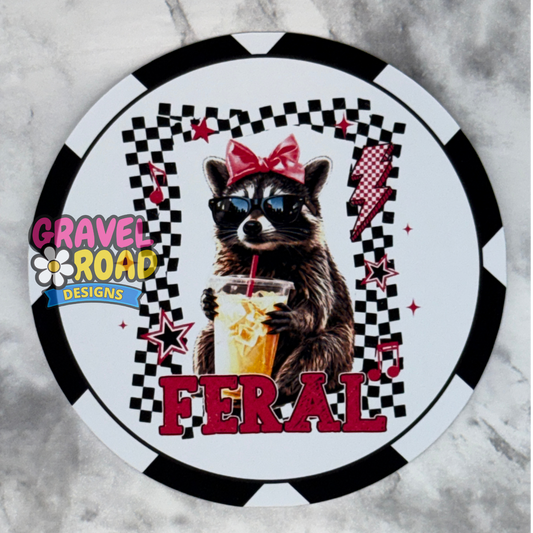 A circular cardstock design featuring a raccoon wearing sunglasses and a red bow, holding an iced drink. The design includes a black and white checkered border, red lightning bolts, stars, and the word 'FERAL' in large red letters.