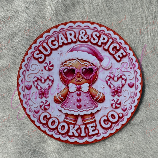 A round cardstock featuring a gingerbread girl in a Santa hat and heart-shaped sunglasses, with the text 'SUGAR & SPICE COOKIE CO'. Displayed on a gray faux-fur background.