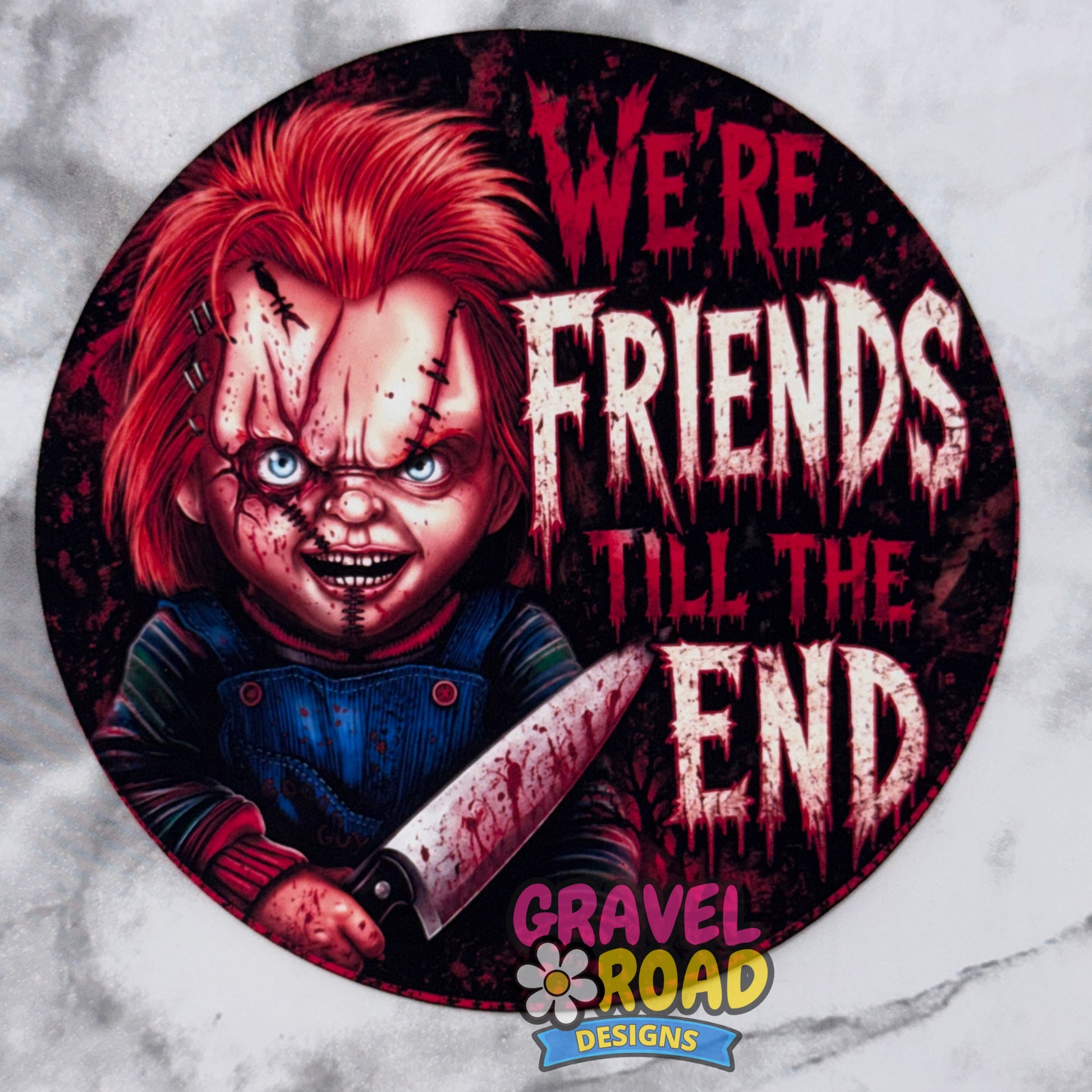 A circular cardstock print featuring a horror character with red hair and blue overalls holding a knife, with the text 'WE'RE FRIENDS Till THE END' in distressed white and red lettering. The design includes a 'GRAVEL ROAD DESIGNS' logo at the bottom. On a grey marble background.