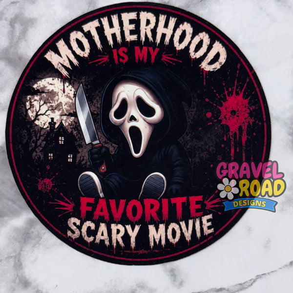 A circular cardstock featuring a Ghostface-style hooded figure holding a knife, with the text 'MOTHERHOOD IS MY FAVORITE SCARY MOVIE' on a dark background. A 'Gravel Road Designs' logo appears in the lower right of the design. On a grey marble background.