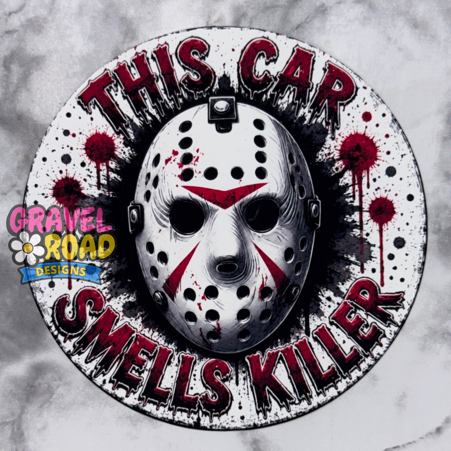 A circular cardstock featuring a hockey mask with red markings, surrounded by the text 'THIS CAR SMELLS KILLER' in distressed red lettering. The design includes blood splatter details and a 'Gravel Road Designs' logo on the left. On a grey marble background.