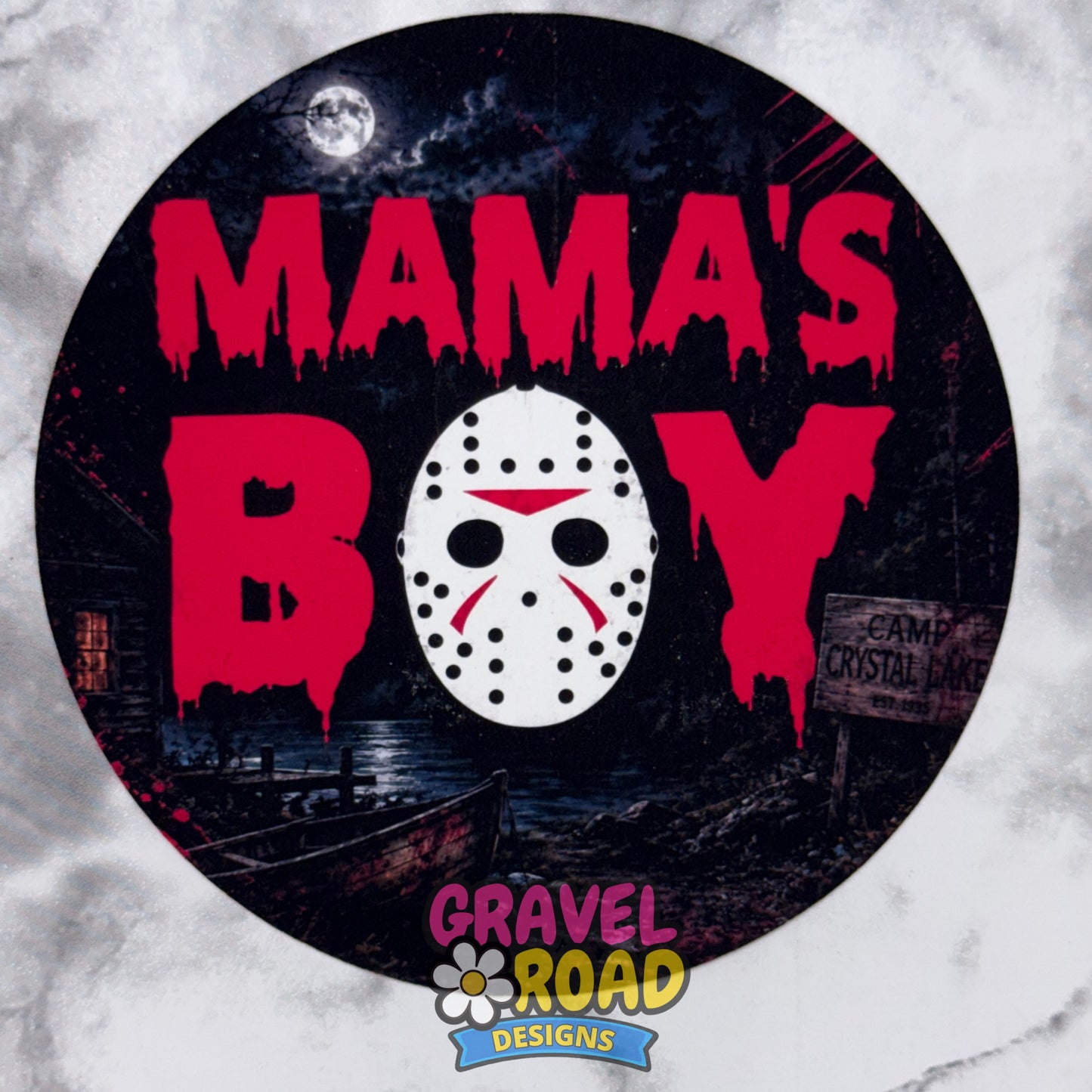 A circular Halloween-themed cardstock featuring the text 'MAMA'S BOY' in dripping red letters, with a white hockey mask in the center. The background depicts a dark, spooky campsite scene with a full moon and a cabin. Gravel Lane Designs logo at the bottom. On a grey marble background.
