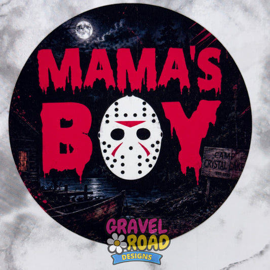 A circular Halloween-themed cardstock featuring the text 'MAMA'S BOY' in dripping red letters, with a white hockey mask in the center. The background depicts a dark, spooky campsite scene with a full moon and a cabin. Gravel Lane Designs logo at the bottom. On a grey marble background.