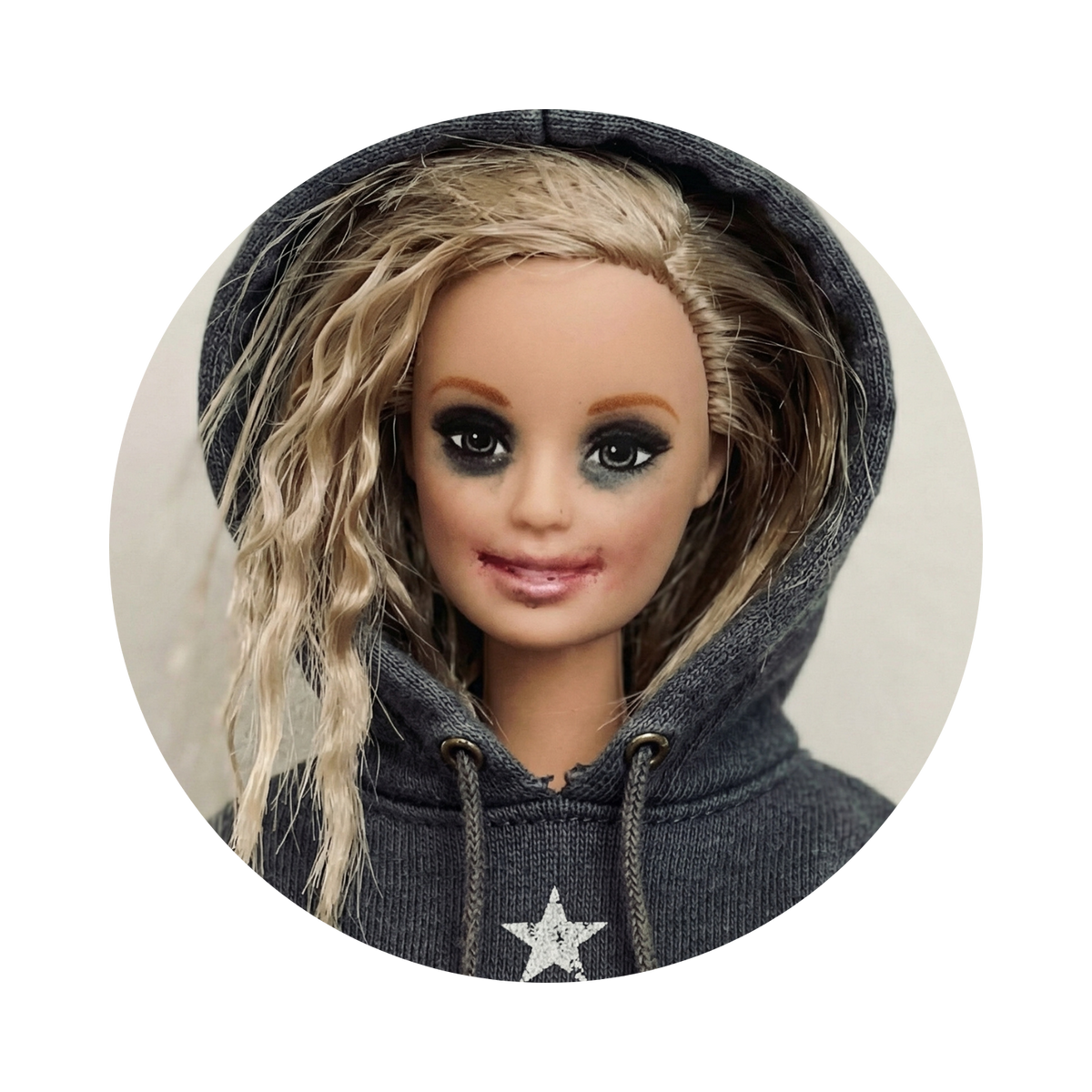A doll with long wavy blonde hair wearing a dark gray hoodie with a silver glittery star on the front, shown inside a circular frame against a light gray background.