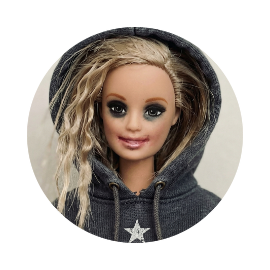 A doll with long wavy blonde hair wearing a dark gray hoodie with a silver glittery star on the front, shown inside a circular frame against a light gray background.