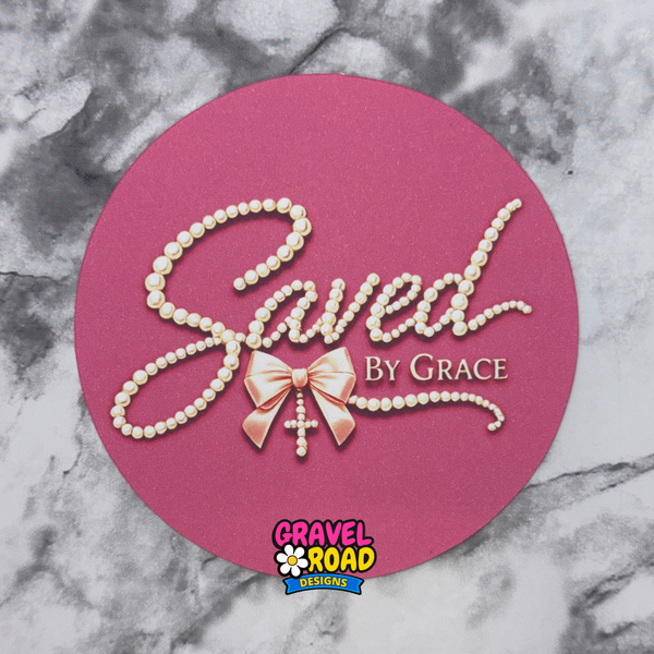 Circular deep pink logo sticker with the words “Saved by Grace” in gold pearl-style lettering. The word “Saved” is written in large cursive script made of bead-like dots, with a gold bow and small cross pendant hanging beneath the center. “BY GRACE” appears in smaller capital letters to the right. The sticker is photographed on a gray and white marble background, with a “Gravel Road Designs” watermark at the bottom.