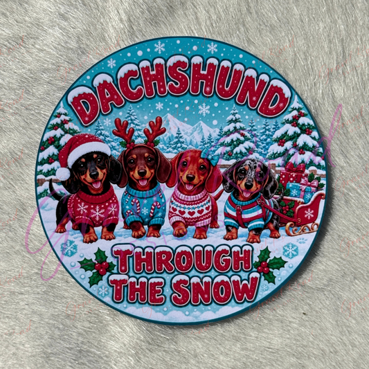 A round printed coaster cardstock featuring four dachshunds dressed in holiday sweaters and hats, standing in a snowy winter scene with the words “DACHSHUND THROUGH THE SNOW” in large red letters. Displayed on a faux fur background.