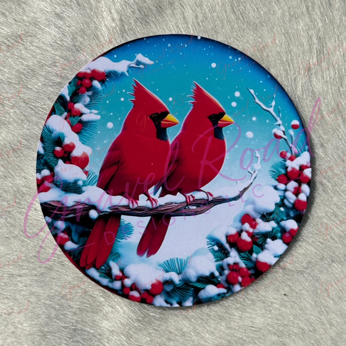 A round cardstock featuring a winter holiday design with two red cardinals perched on snowy branches surrounded by red berries against a blue sky background, displayed on a gray textured surface.