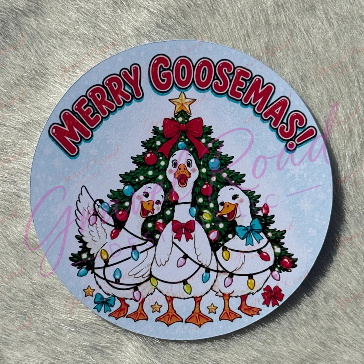 A round printed cardstock featuring three white cartoon geese around a decorated Christmas tree, with the text “MERRY GOOSEMAS!” in red arched letters on a pale blue background. Displayed on a faux fur background.