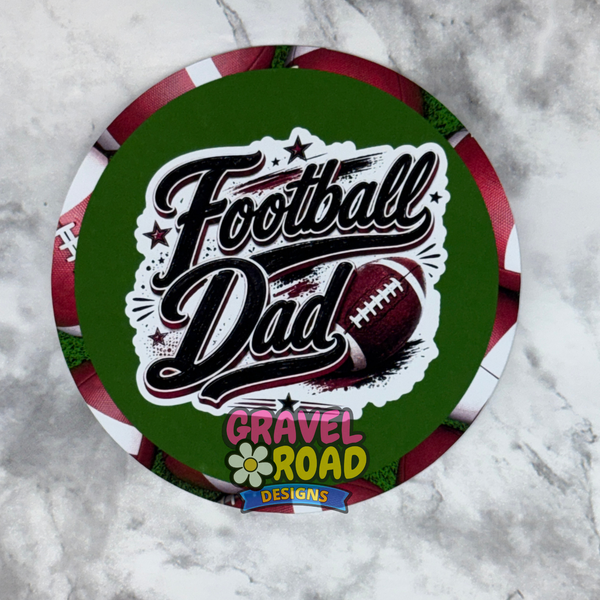 A round cardstock with a football theme, featuring the words 'Football Dad' in stylized black script over a green background, with a football graphic and the logo name 'Gravel Road Designs' at the bottom. The sticker is placed on a light gray marble surface.