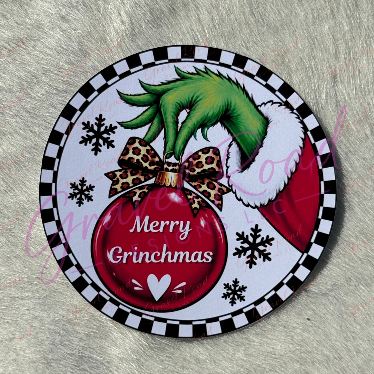 A round printed cardstock featuring a Christmas design, with a green hand holding a red ornament and bow, on a furry gray surface.