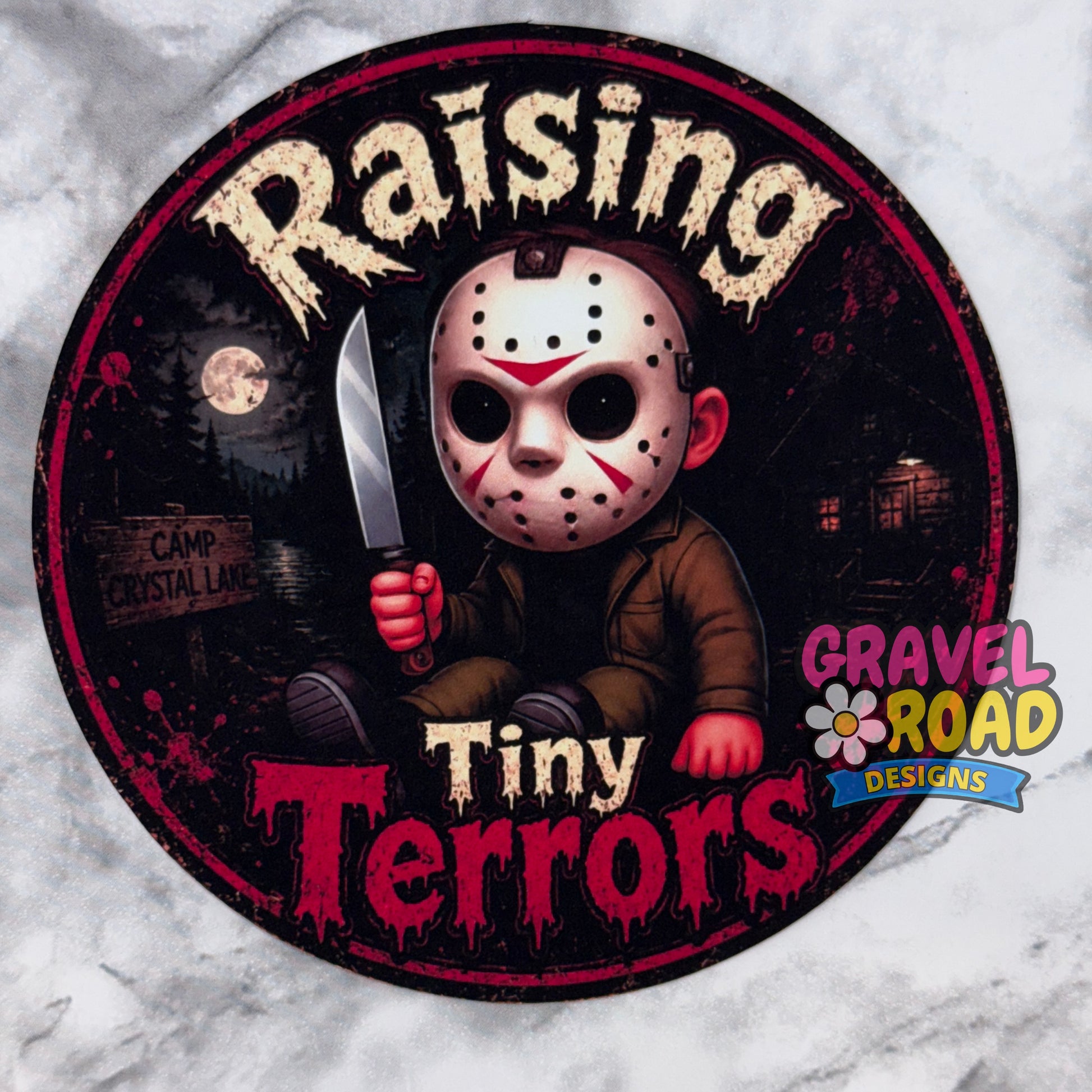 A circular cardstock featuring a stylized horror character in a hockey mask holding a knife, with the text 'Raising Tiny Terrors' and a 'Gravel Road Designs' logo, shown on a light marble-like background.