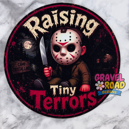 A circular cardstock featuring a stylized horror character in a hockey mask holding a knife, with the text 'Raising Tiny Terrors' and a 'Gravel Road Designs' logo, shown on a light marble-like background.