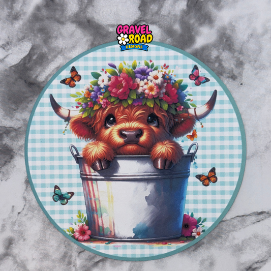 Circular design with a cartoon cow in a bucket, flowers, and butterflies on a checkered background. Gravel Road Designs logo visible.