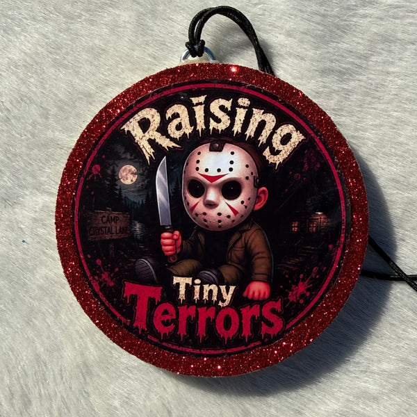 Cartoon child horror‑themed freshie with a glittery red border, black hanging cord, and “Raising Tiny Terrors” text in front of a moonlit forest and camp sign