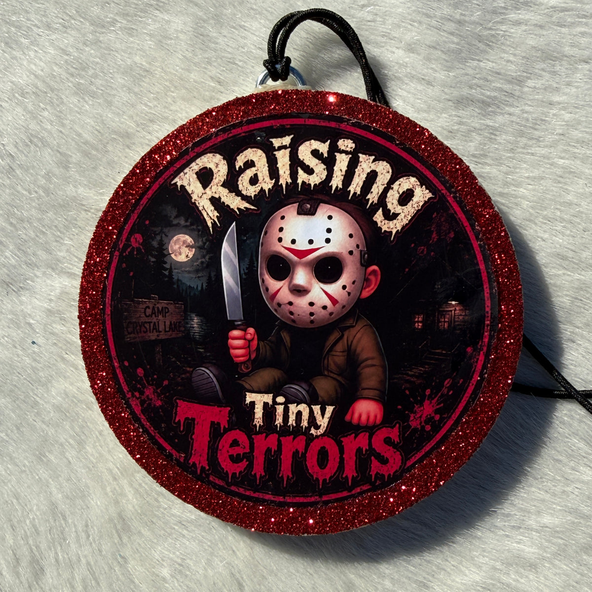 Cartoon child horror‑themed freshie with a glittery red border, black hanging cord, and “Raising Tiny Terrors” text in front of a moonlit forest and camp sign