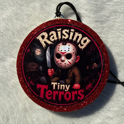 Cartoon child horror‑themed freshie with a glittery red border, black hanging cord, and “Raising Tiny Terrors” text in front of a moonlit forest and camp sign