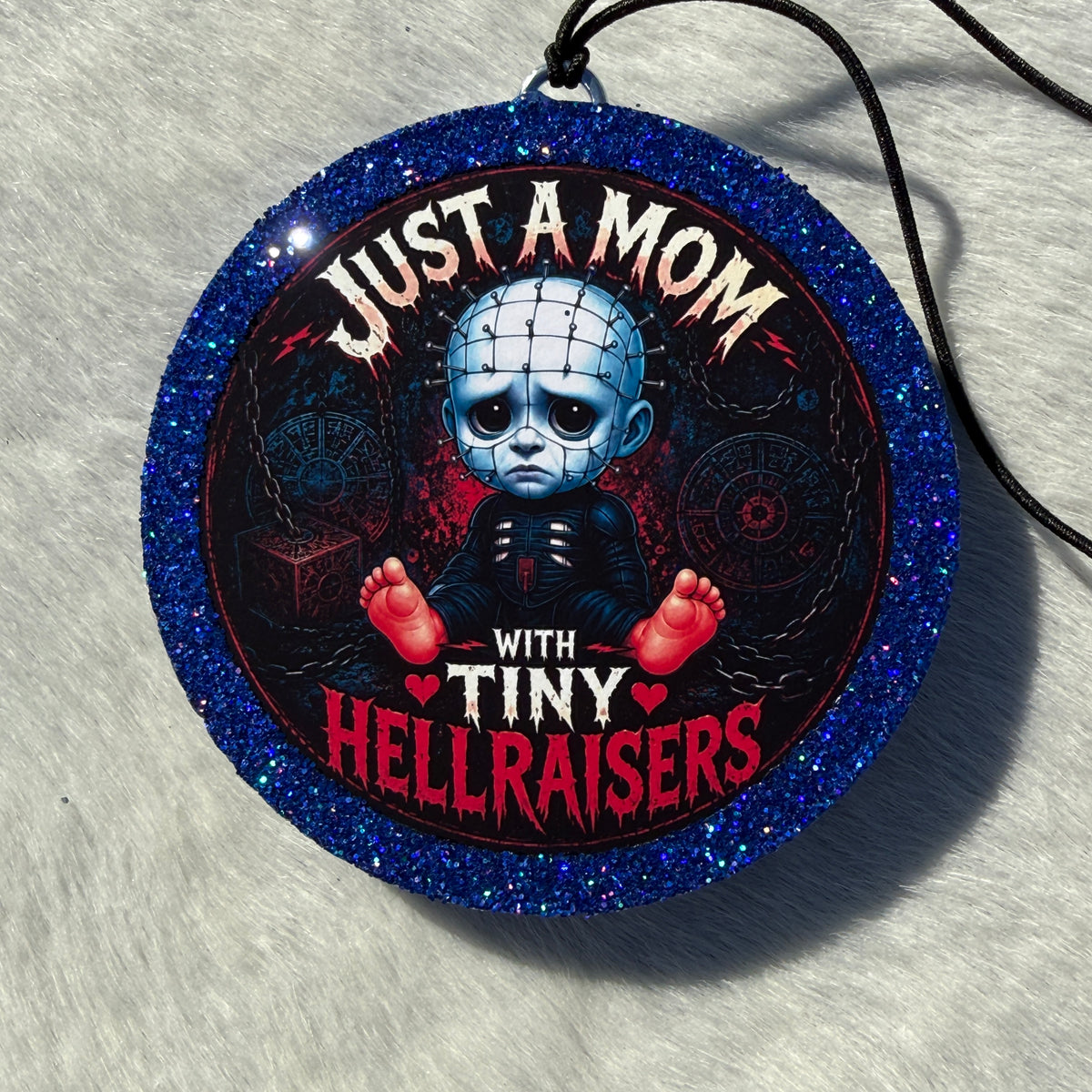 Cartoon baby horror‑themed freshie with a glittery blue border, black hanging cord, and “Just a Mom With Tiny Hellraisers” text on a dark chain‑themed background.