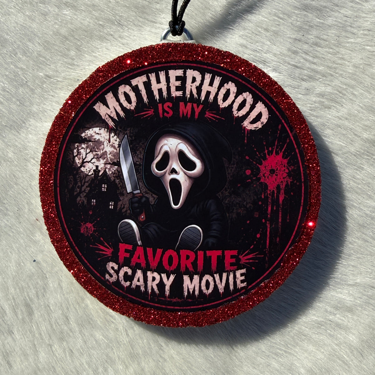 A round glittery red hanging car freshie featuring a Ghostface-style horror character and the text 'MOTHERHOOD IS MY FAVORITE SCARY MOVIE' on a white textured background.